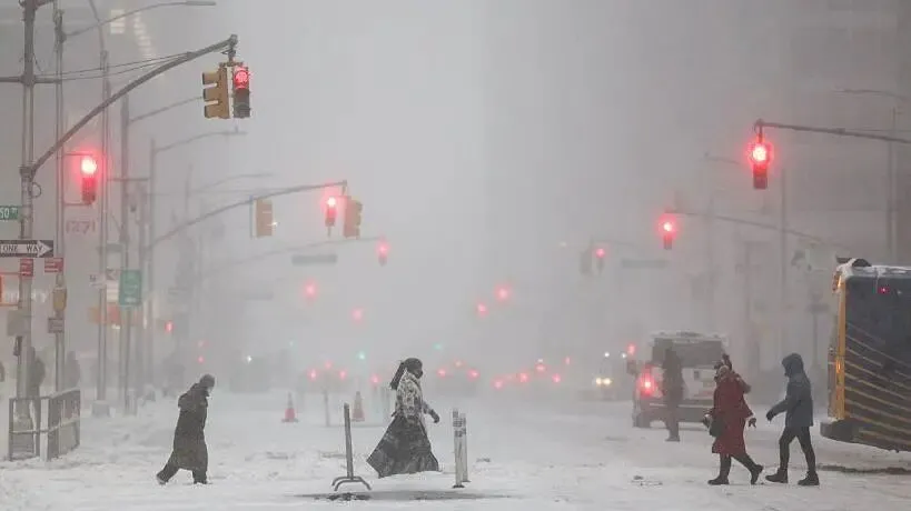 People walking across Sixth Avenue as snow falls. There's snow on the ground and lots of traffic lights on red. Cars are in the distance with their brake lights on.