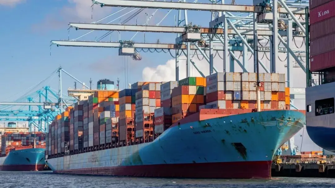 Dozens of multi-colored containers are stacked on a blue cargo ship docked at a port under cranes. The cargo ship is between two other vessels.