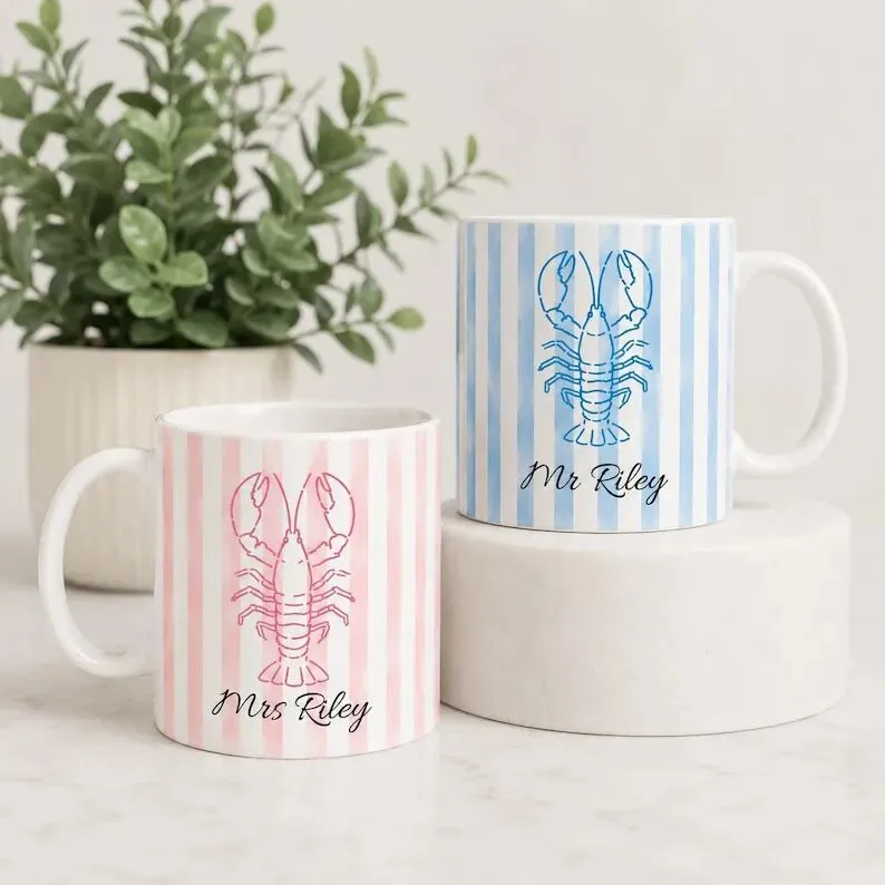 May include: Two ceramic mugs featuring vertical stripes and a central lobster illustration. One mug has pink stripes with the text Mrs Riley, while the other has blue stripes with the text Mr Riley. Both mugs have a handle and a cylindrical shape.